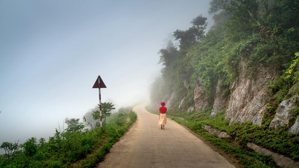 Woman in a red top and cream skirt walking alone on a foggy mountain road surrounded by lush greenery—symbolizing slow travel and mindful exploration