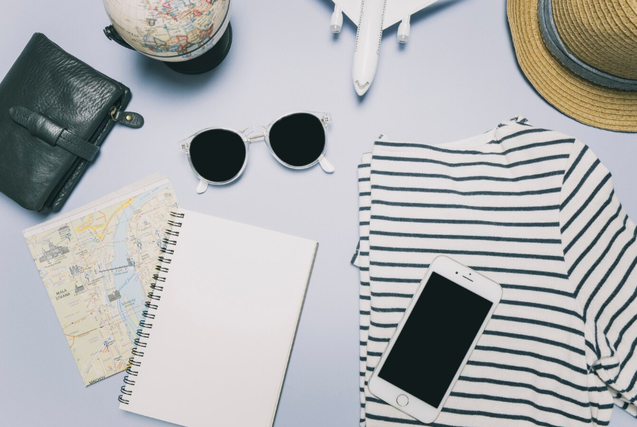 Flat lay of minimalist travel essentials including striped shirt, map, notebook, sunglasses, and phone