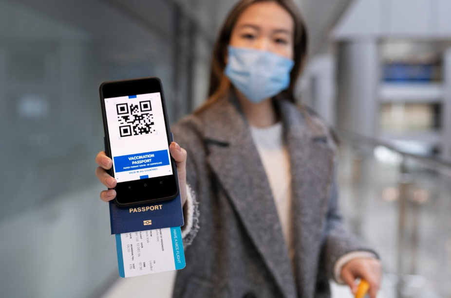 Woman at airport wearing a mask, holding a smartphone with a digital vaccination passport, passport, and boarding pass