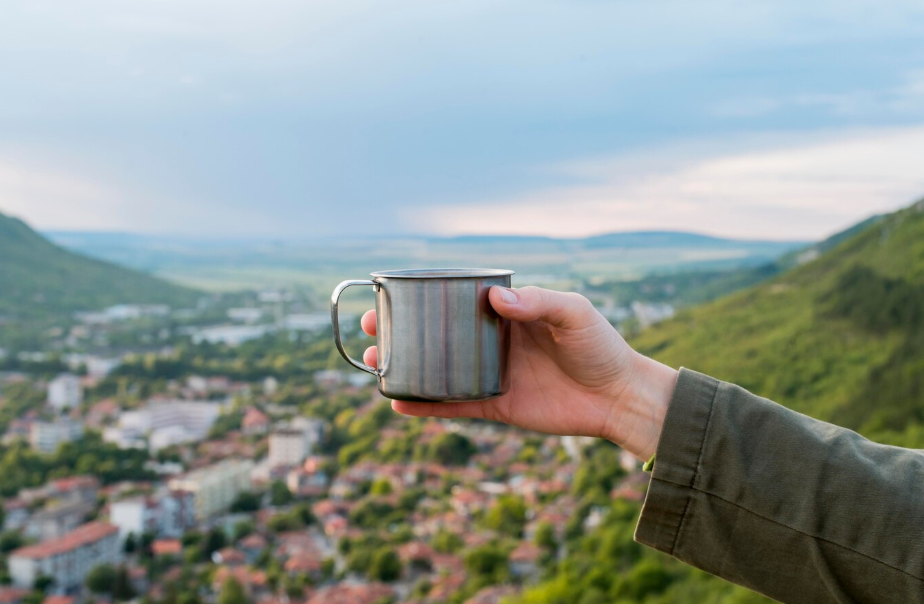 Traveler holding a metal mug overlooking a scenic town from the hills.