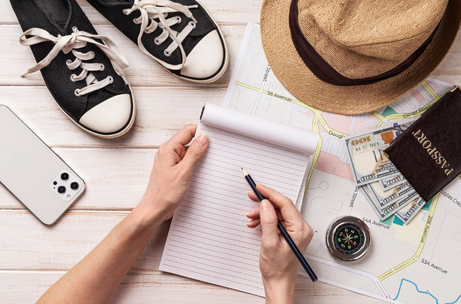 Travel essentials checklist flat lay with map, sneakers, hat, passport, cash, smartphone, compass, and a person writing a packing list.