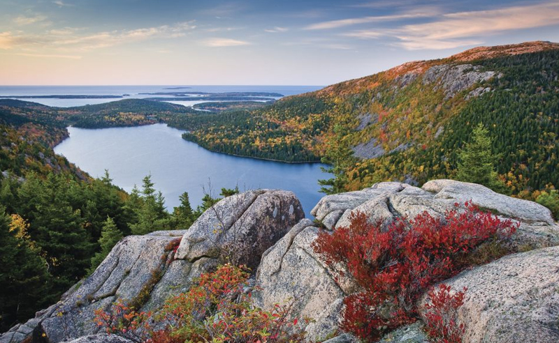 Scenic overlook of Jordan Pond and fall foliage in Acadia National Park, featuring rocky cliffs and colorful trees.