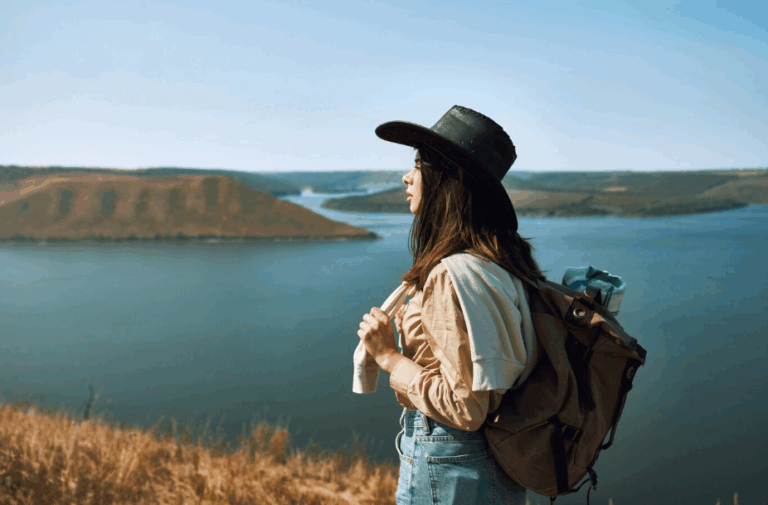 Female solo traveler wearing a wide-brimmed hat and backpack, standing on a hillside overlooking a lake