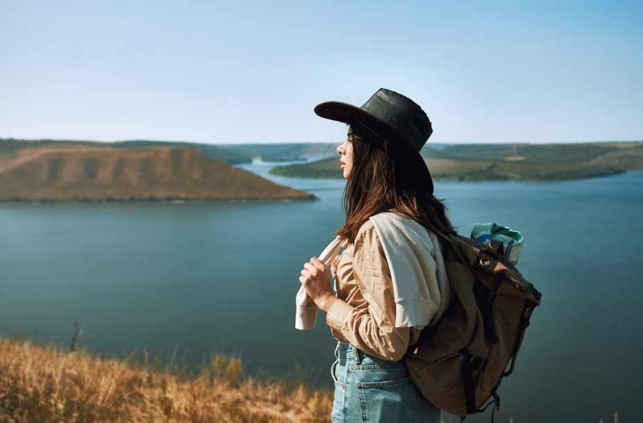 Female solo traveler wearing a wide-brimmed hat and backpack, standing on a hillside overlooking a lake