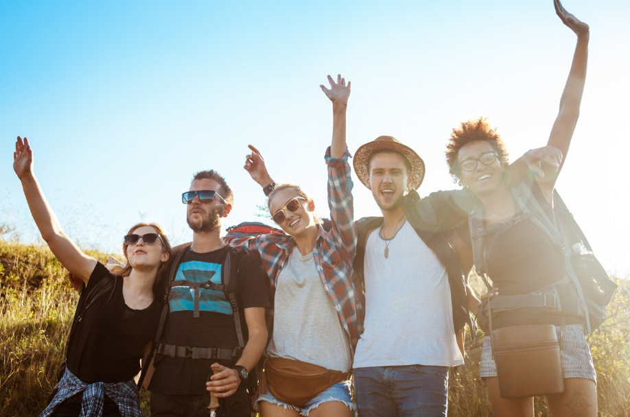 Group of young friends hiking outdoors on a sunny day, smiling and raising their hands.